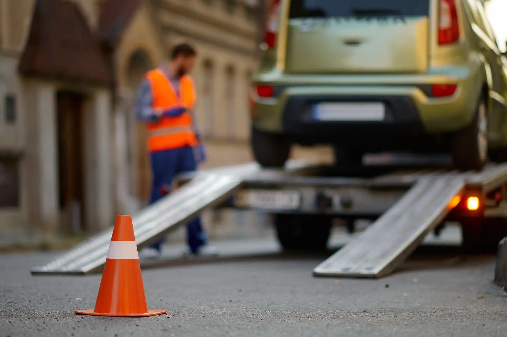 pomoc drogowa, pracownik w pomarańczowej kamizelce odblaskowej sprawdza ustawienie samochodu osobowego na lawecie; z tyłu lawety wysunięte podjazdy przed którymi stoi pachołek