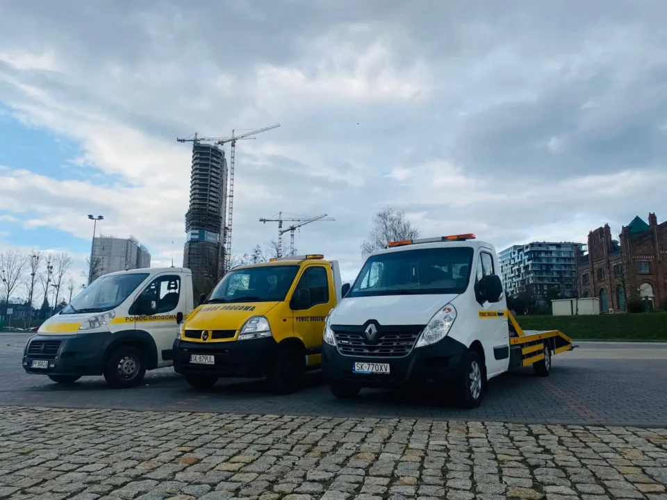 trzy samochody laweta pomocy drogowej Katowice, zaparkowane obok siebie, pochmurny dzień, parking z kostki brukowej, w tle dźwig budowlany i wieżowiec w trakcie budowy