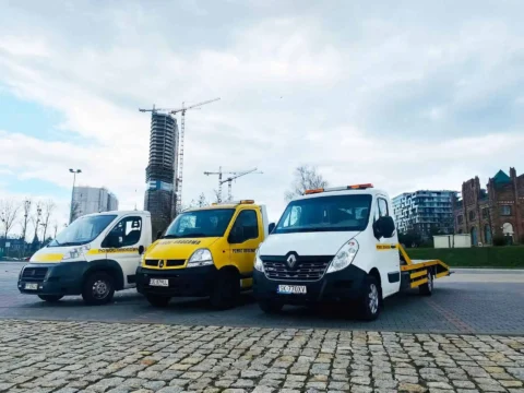 trzy samochody laweta pomocy drogowej Katowice, zaparkowane obok siebie, pochmurny dzień, parking z kostki brukowej, w tle dźwig budowlany i wieżowiec w trakcie budowy